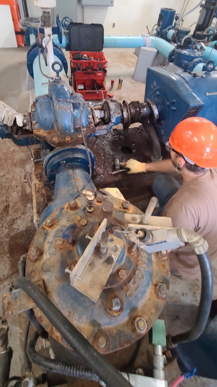 A worker wearing an orange hard hat and gloves repairing or maintaining industrial equipment with rusted parts and large pipes in a factory setting.