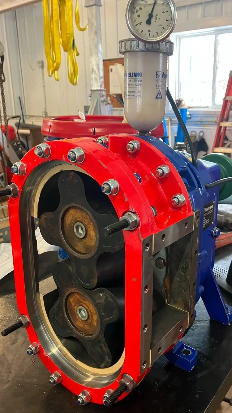 Close-up of a mechanical engine with a red and blue casing, partially disassembled, including metal components and a pressure gauge on top.