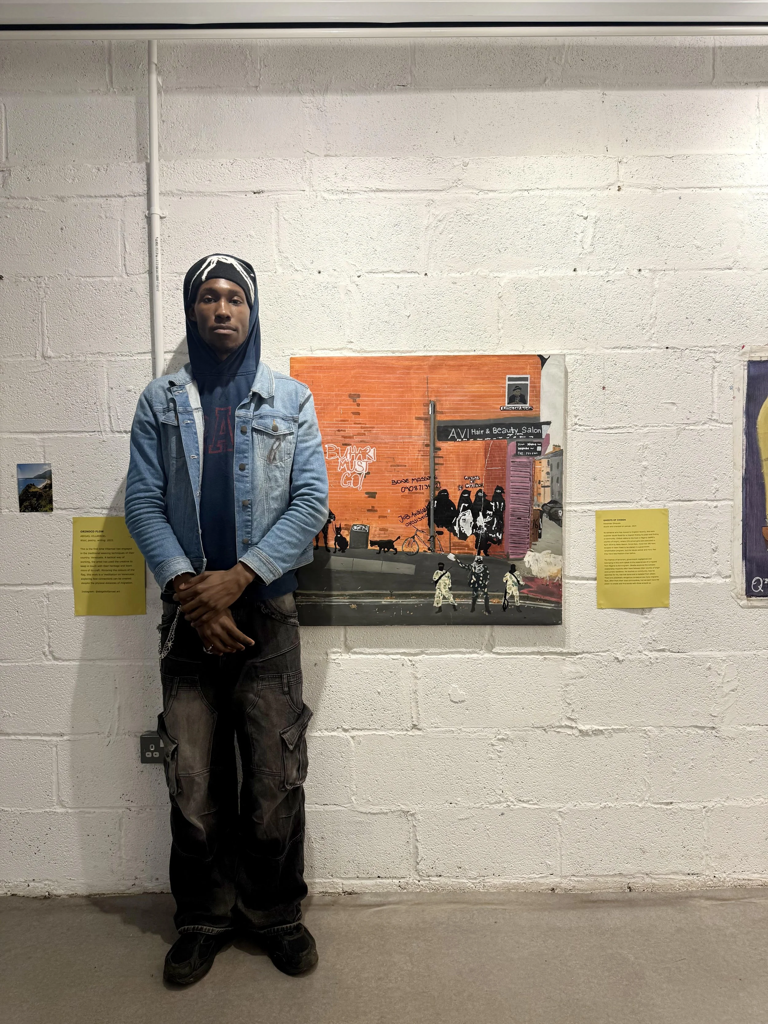 A young man standing in an art gallery with his arms crossed. He is wearing a denim jacket, hoodie, cargo pants, and sneakers, in front of a wall with artwork and informational plaques.