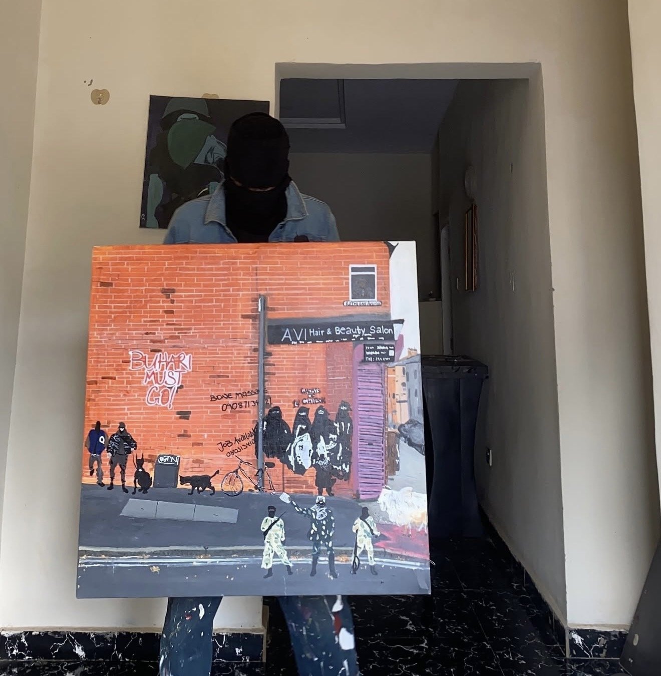 A person with a black face covering and a denim jacket holding a painting of a street scene with graffiti, street art, pedestrians, and bicycles in an indoor setting.