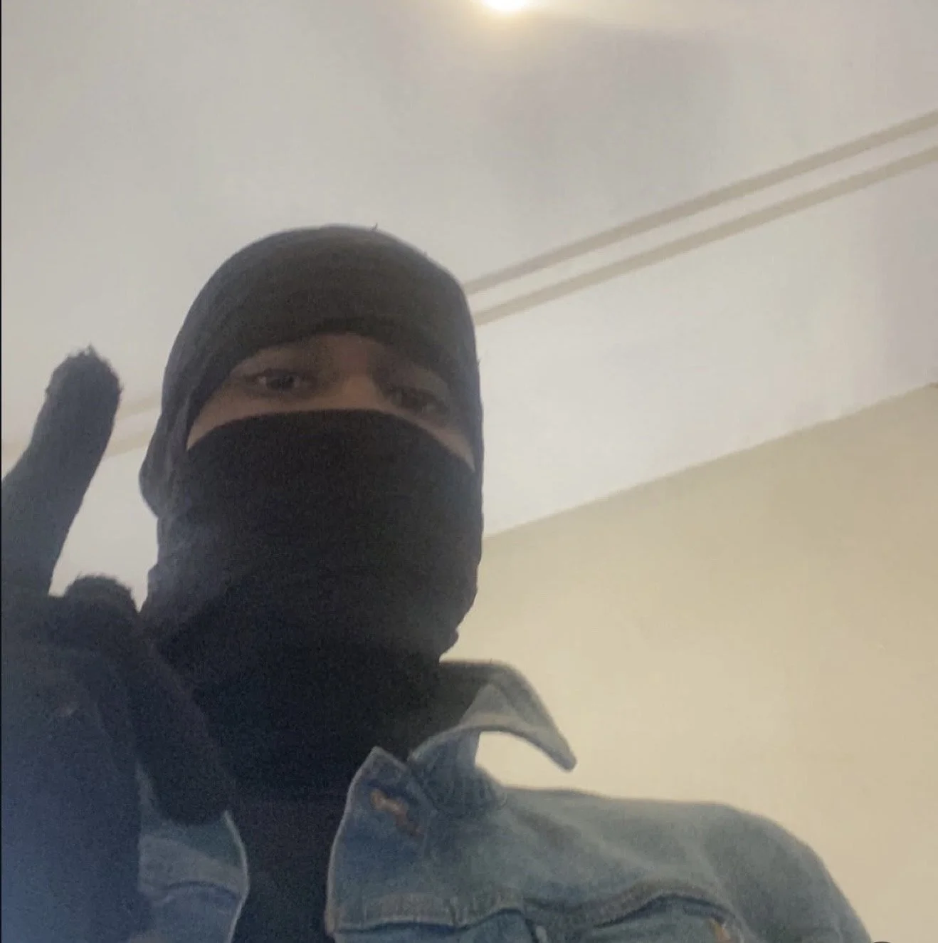 A person wearing a black ski mask, black gloves, and a denim jacket taking a selfie inside a room.