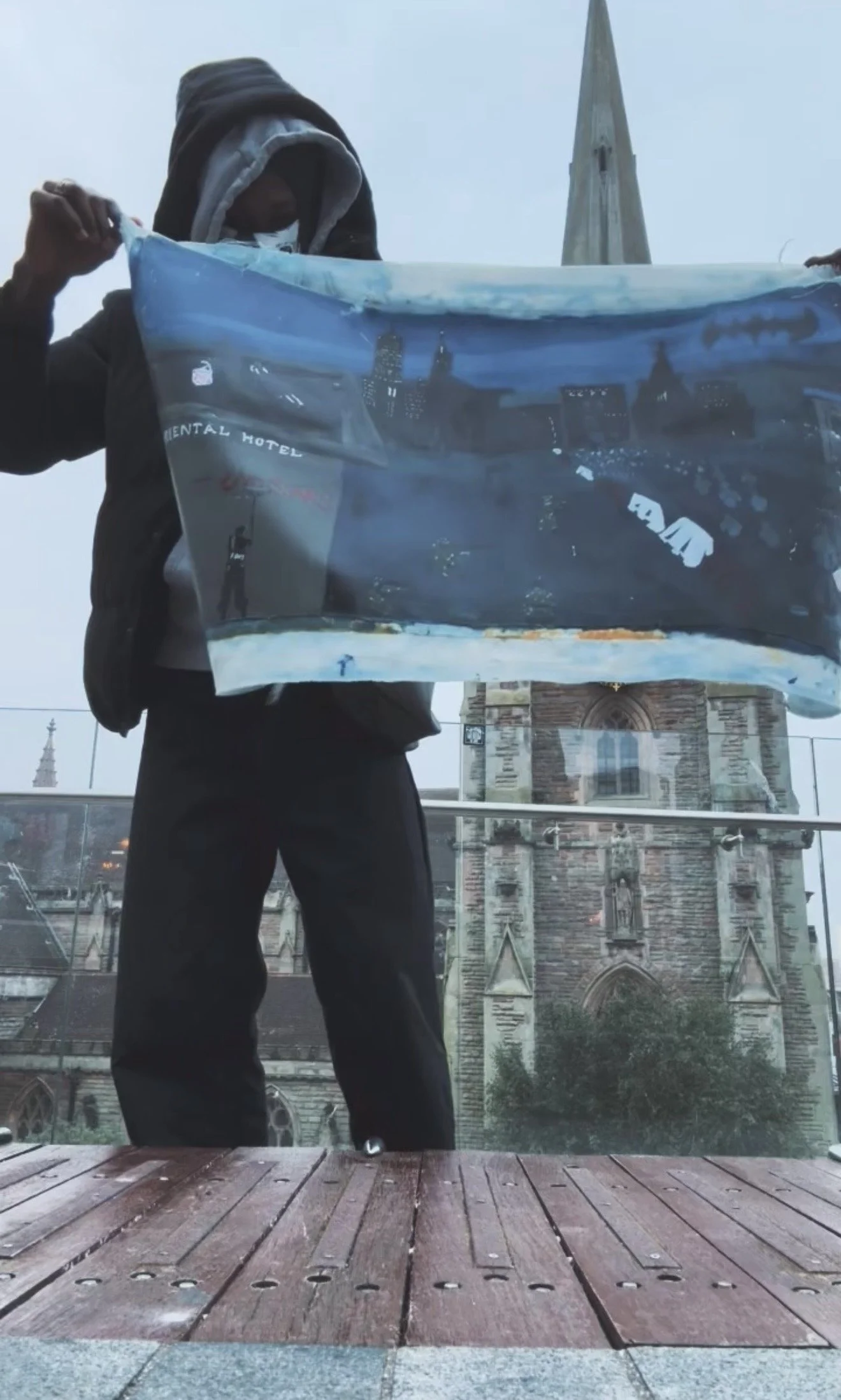 Person holding a poster or sign with a cityscape and church in the background, on a balcony or terrace with wooden flooring.
