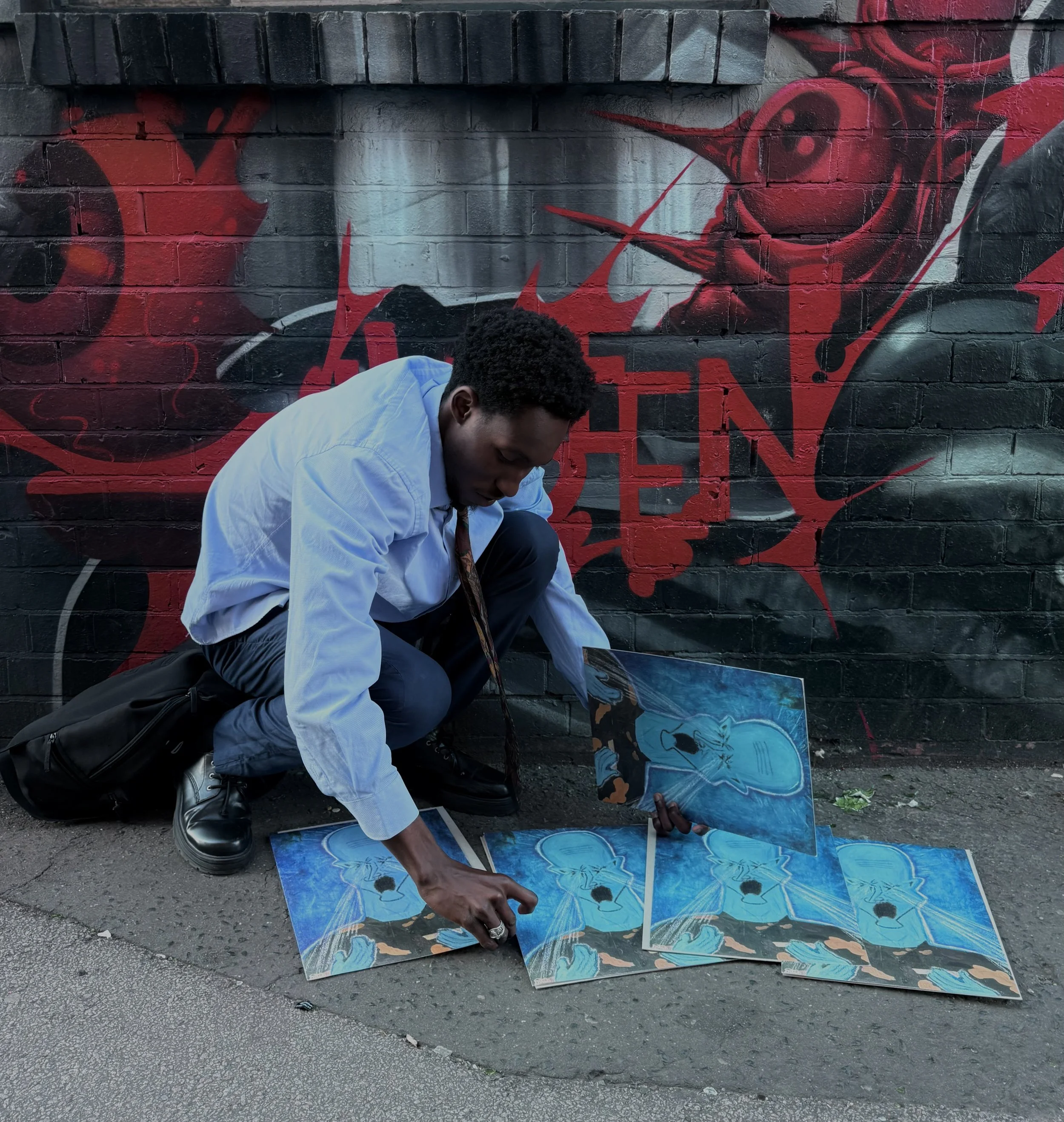 Person with dark curly hair painting graffiti on a brick wall, wearing a light blue shirt and black shoes, with graffiti art in blue, black, red, and white colors in the background.