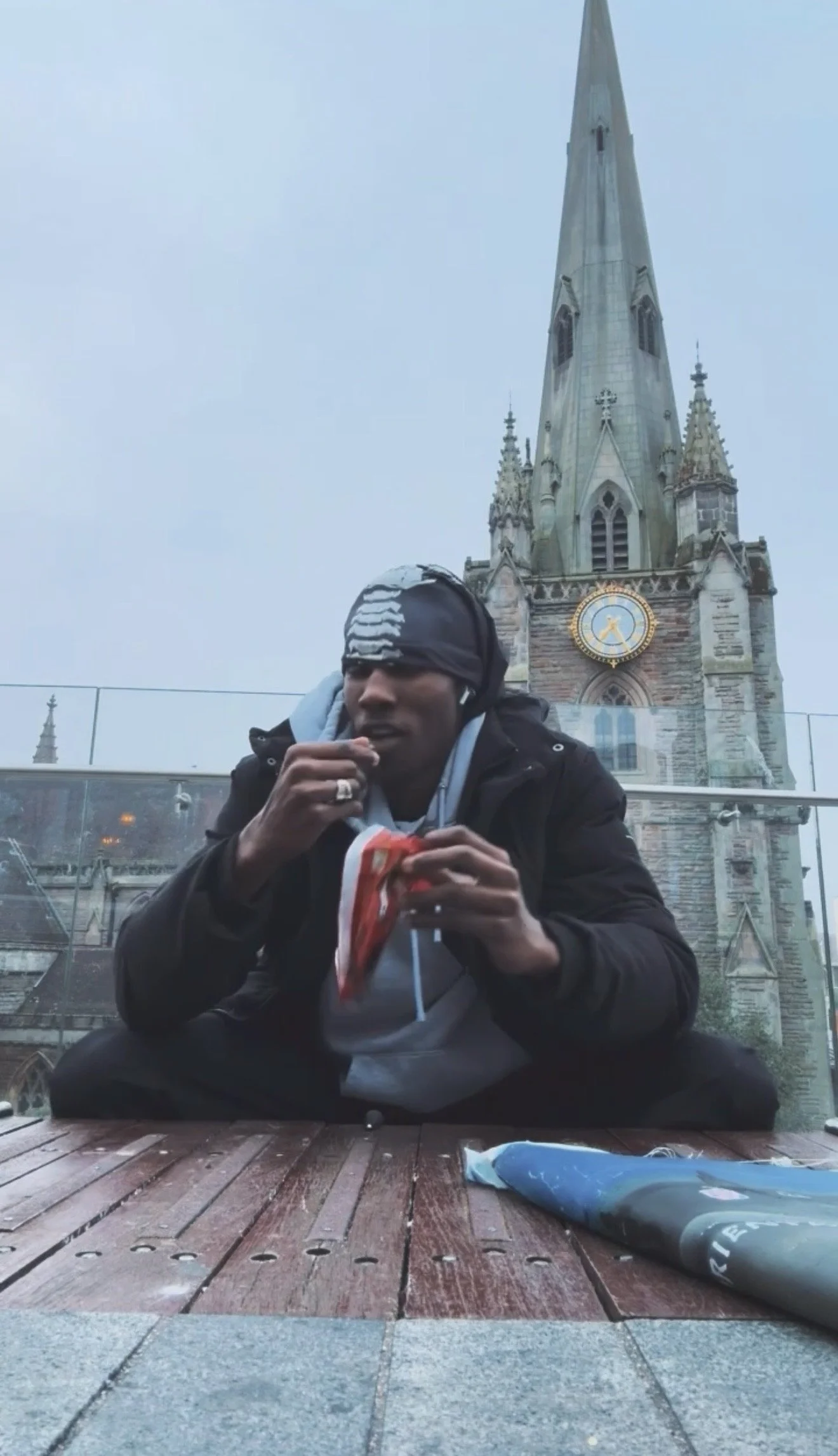 A man sitting on a wooden deck eating snacks, with a historic church with a tall spire and clock in the background.