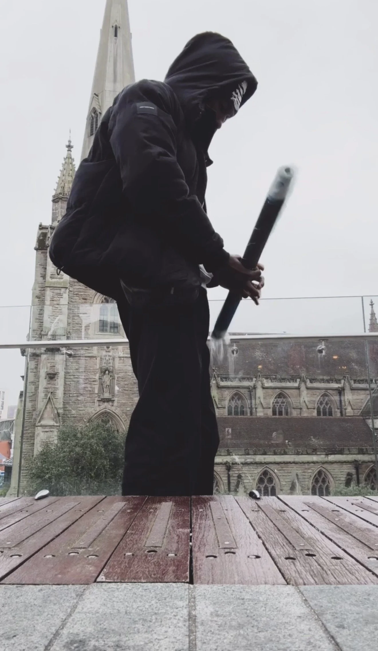 Person with dark clothing and a hood holding what looks like a bat or stick, standing outdoors near a church with tall steeples and stained glass windows, on a cloudy day.