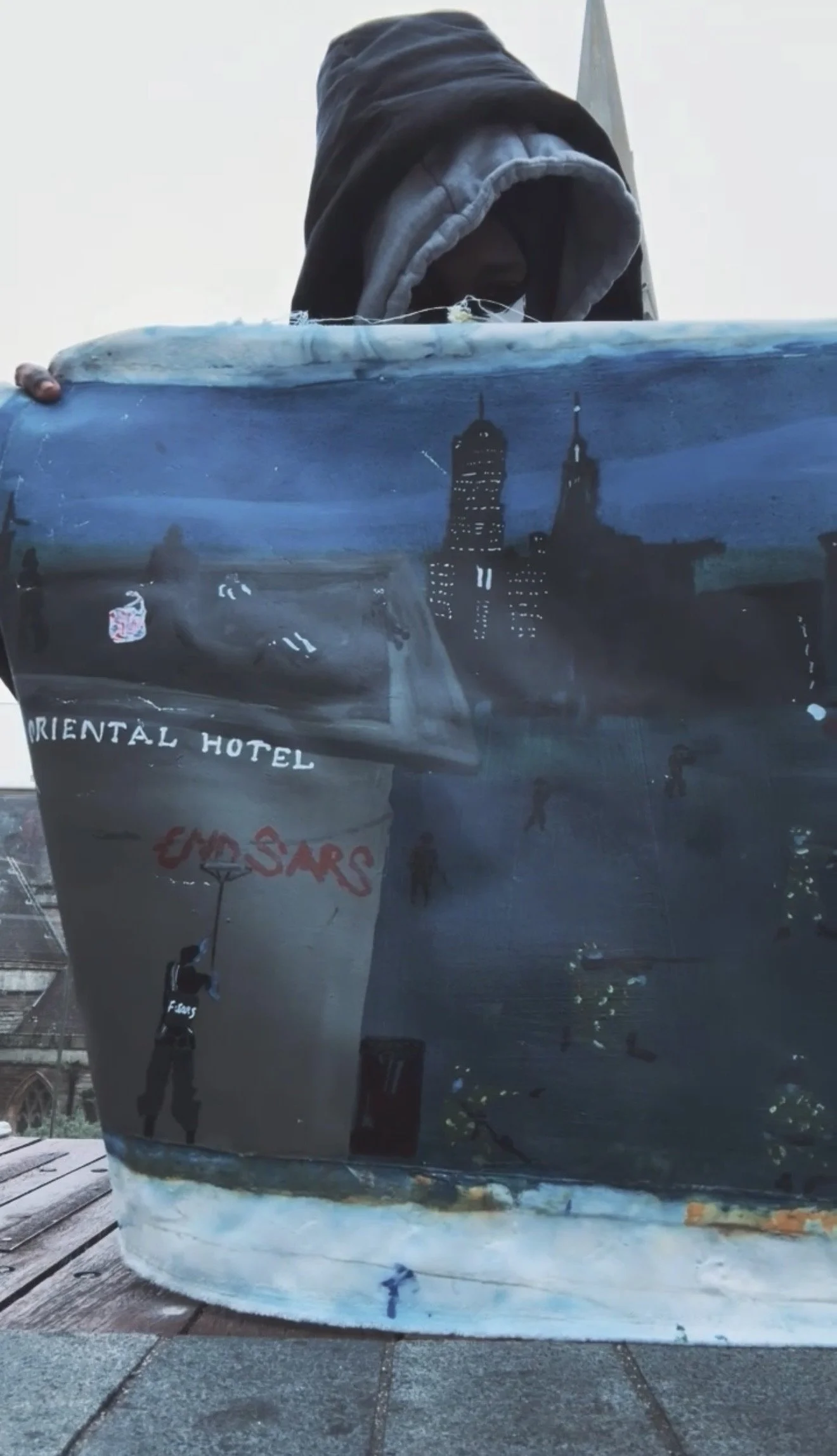 Painting of a cityscape featuring tall skyscrapers and a dark sky, on a container held by a person wearing a hoodie.