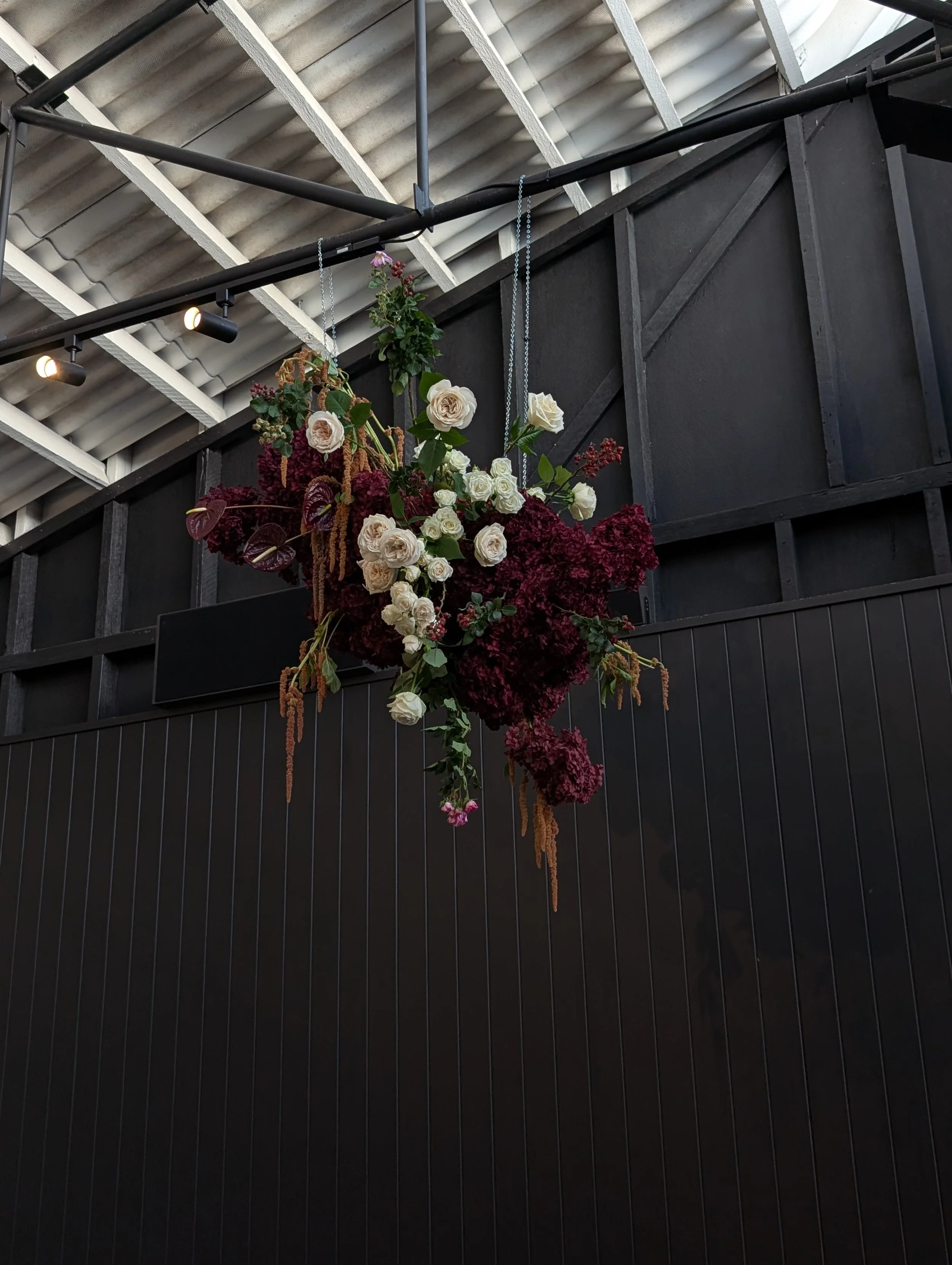 Hanging floral arrangement with white roses, red and maroon flowers, and green leaves, suspended from the ceiling in an indoor setting with black walls and spotlights.