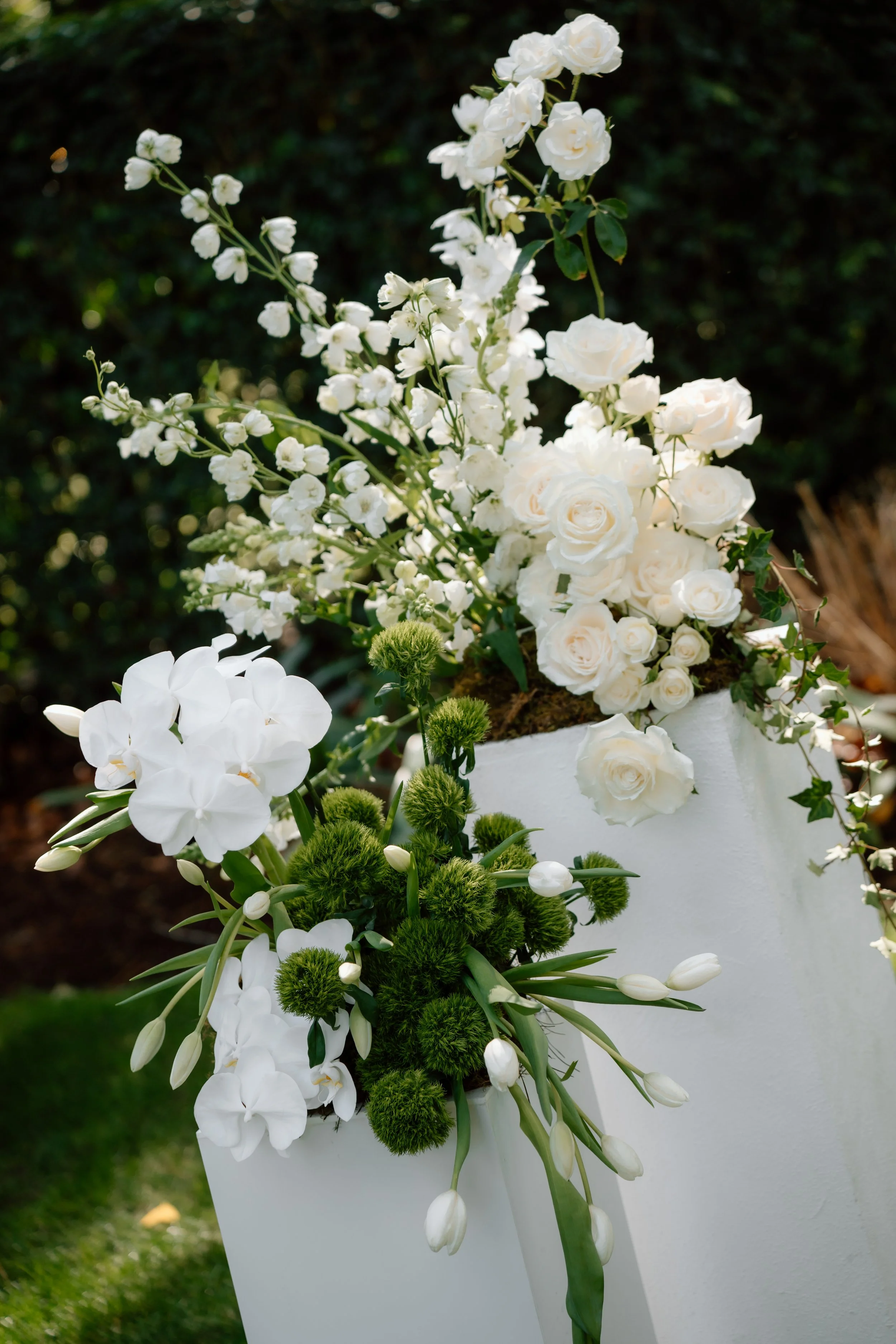 White floral arrangement with various flowers including roses, orchids, and greenery in a white container, outdoors against a blurred green background.