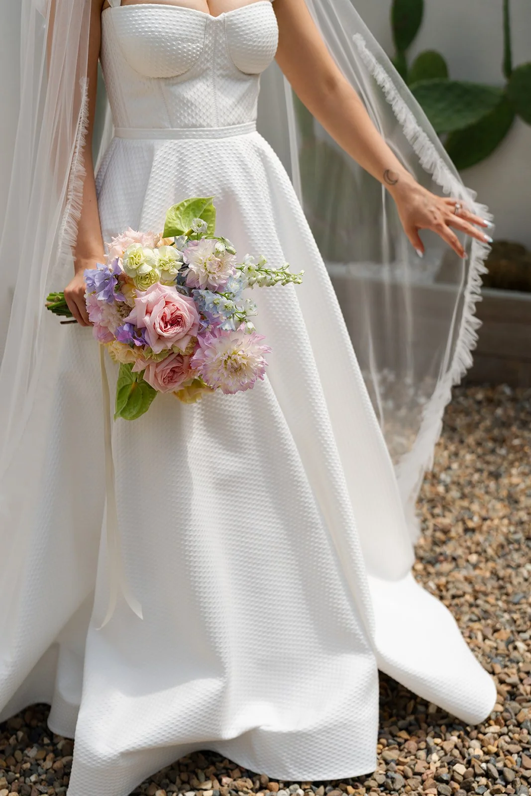 A bride in a white textured wedding dress holding a pastel-colored bouquet of flowers, with her hand extended and a sheer veil draped over her shoulder, standing on a gravel surface.