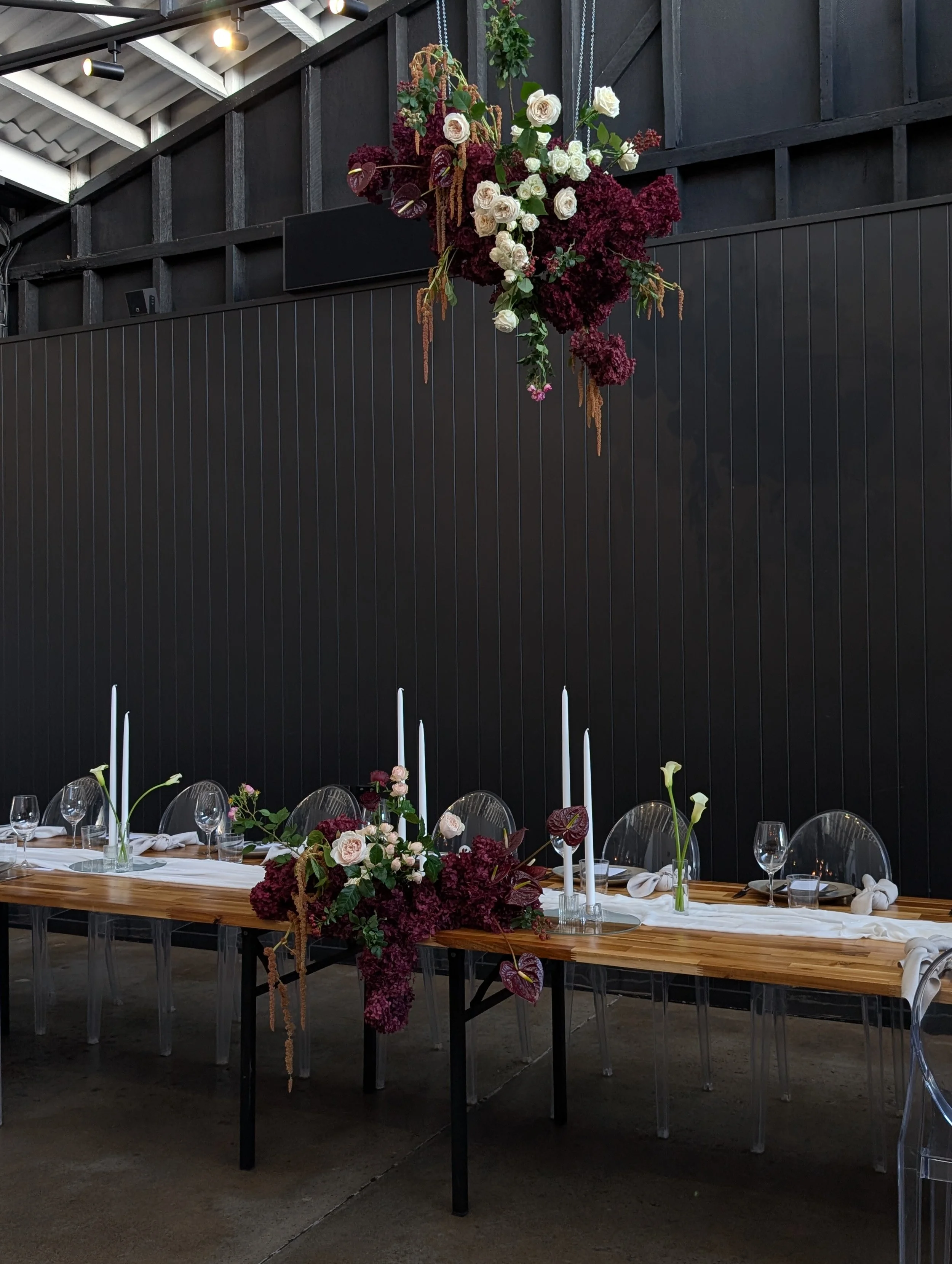 A long wooden table decorated with floral arrangements, white candles, glassware, and white napkins, set for a formal event in an industrial-style venue with black walls and a high ceiling.
