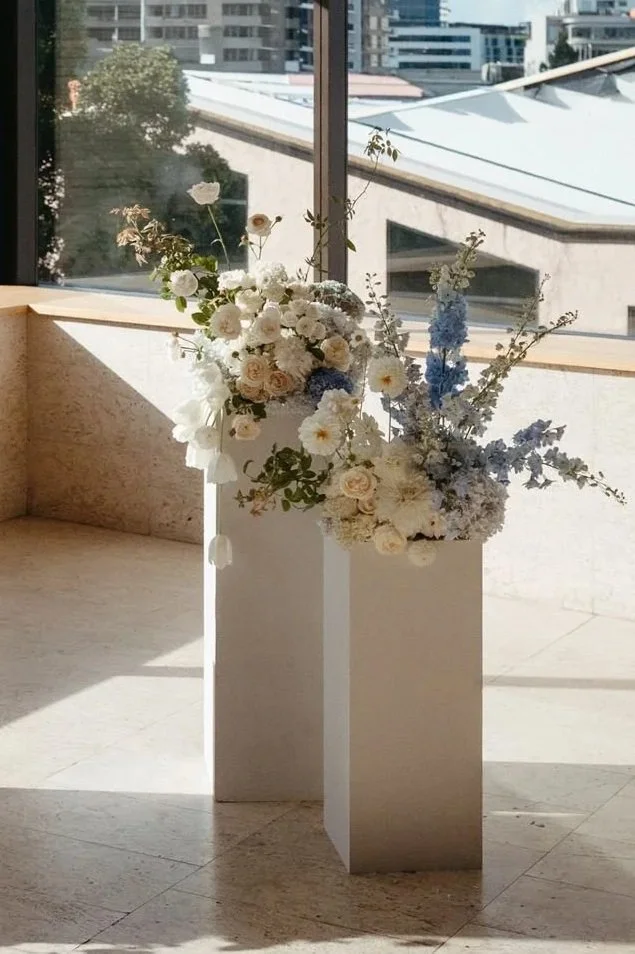 A white vase and a white rectangular pedestal with a bouquet of white and light pink flowers, along with blue delphinium, arranged inside, near a large window in a modern building with cityscape in the background.