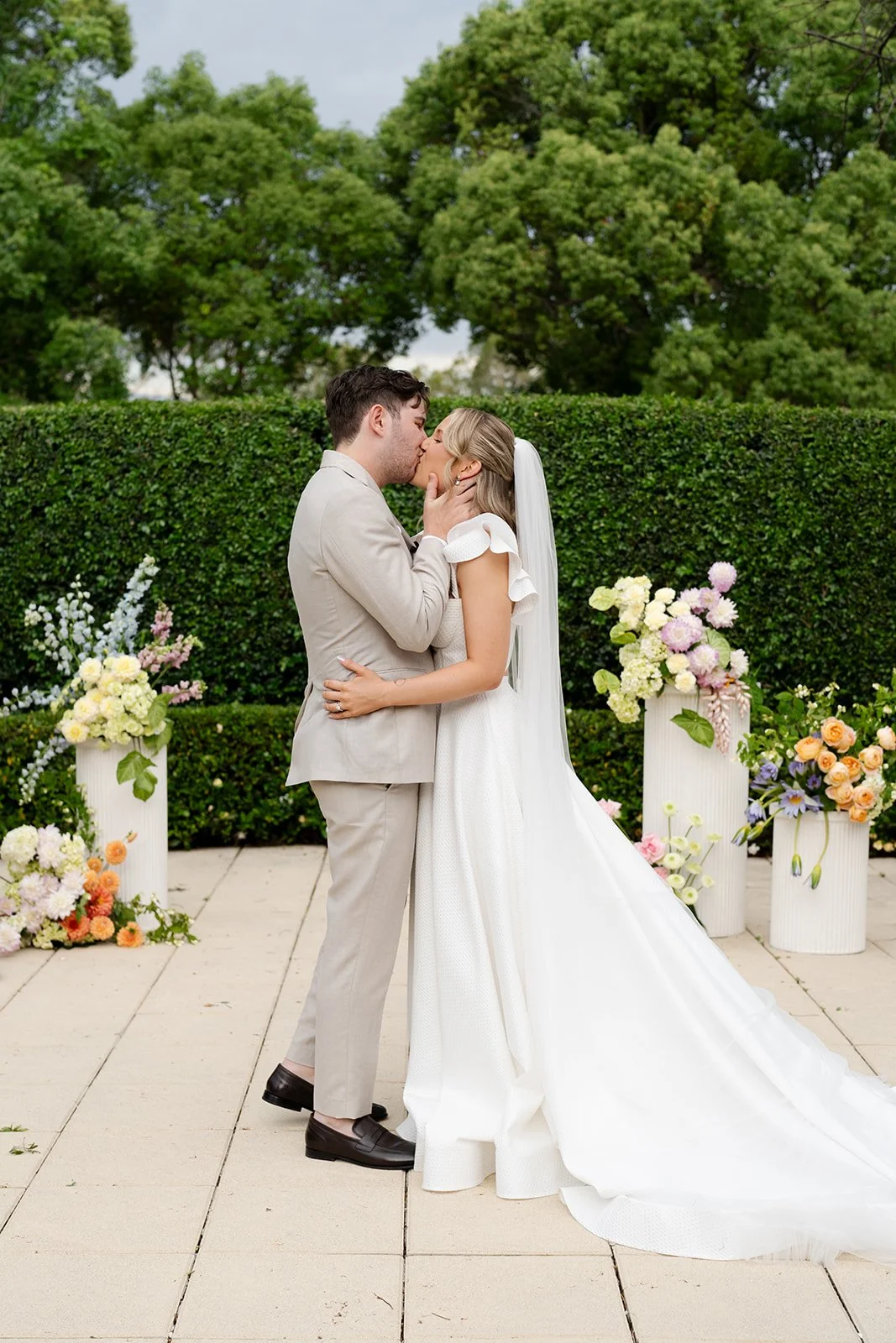 A newlywed couple sharing a kiss outdoors, surrounded by flower arrangements and greenery.