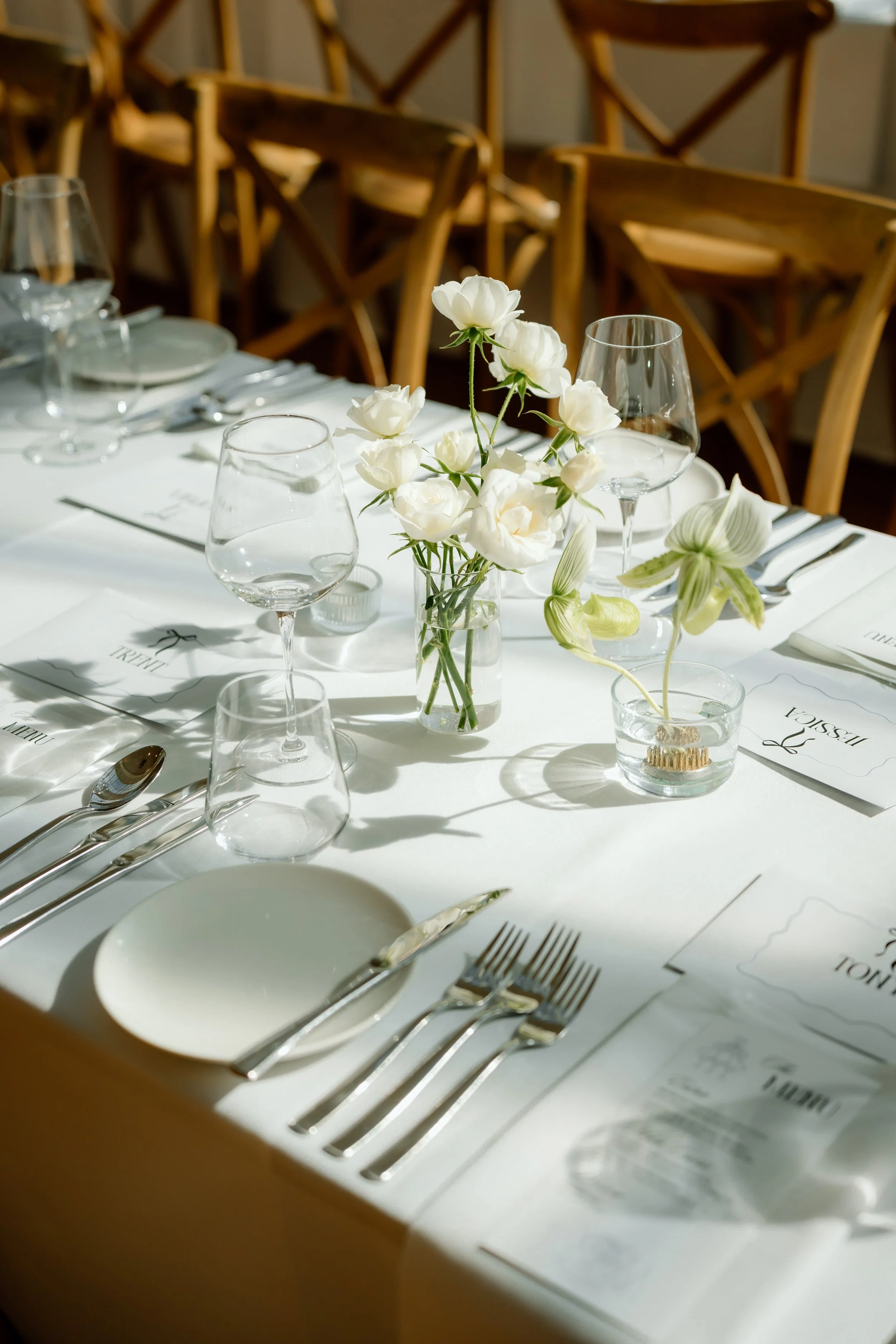 A table set for a formal meal with white tablecloth, glassware, silverware, plates, and a floral centerpiece of white flowers in a glass vase. Wooden chairs are in the background.