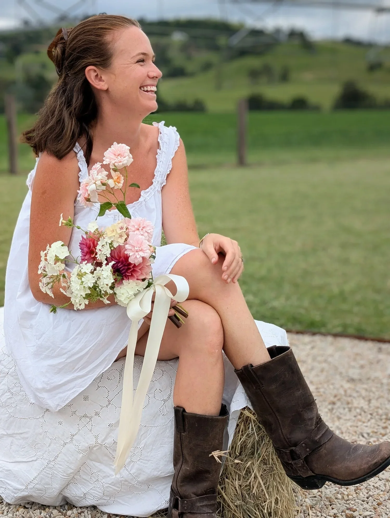 A woman sitting on a baling of hay outdoors, wearing a white dress and brown cowboy boots, holding a bouquet of pink and white flowers, smiling with her eyes closed, with green fields and fencing in the background.