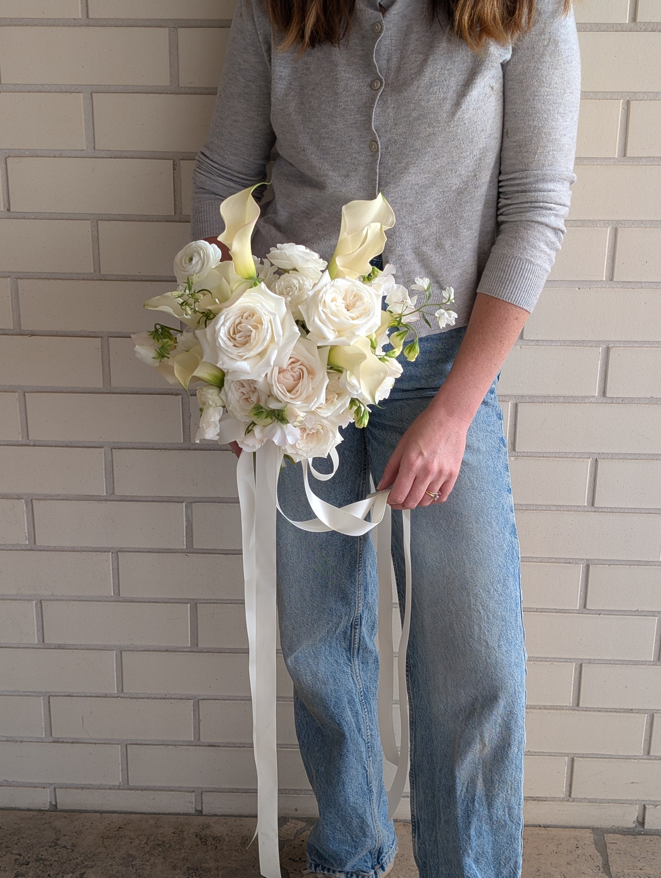 A woman in a gray cardigan and jeans holding a bouquet of white flowers against a brick wall background.