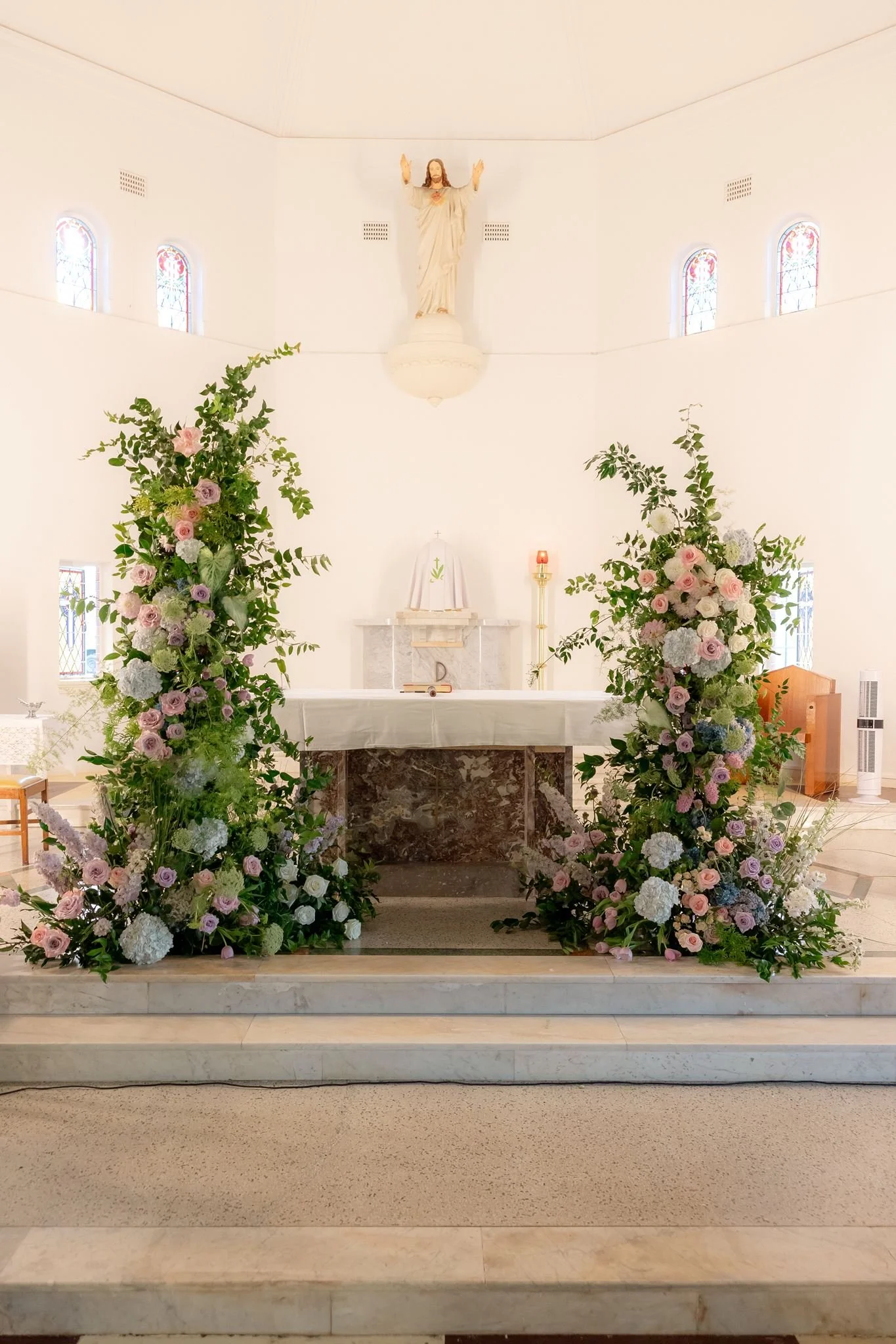 An altar decorated with two large arrangements of pink, white, and purple flowers, with a statue of Jesus Christ above, and a cloth with a cross on it on the altar.