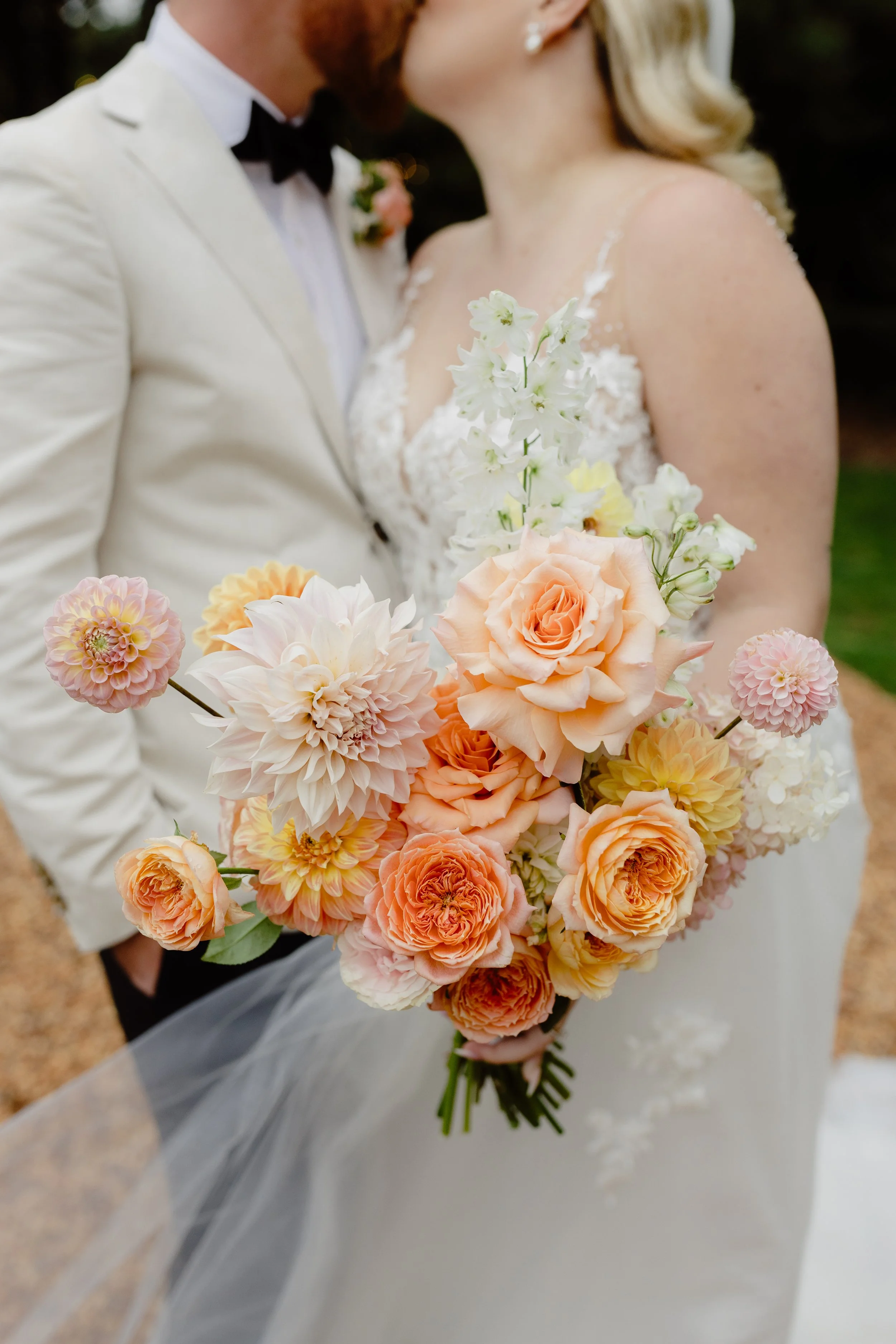 Bride and groom kissing on their wedding day, with the bride holding a large bouquet of peach, white, and pink flowers. The groom is dressed in a white suit jacket with a black bow tie, and the bride is wearing a white lace wedding gown with spaghetti straps.