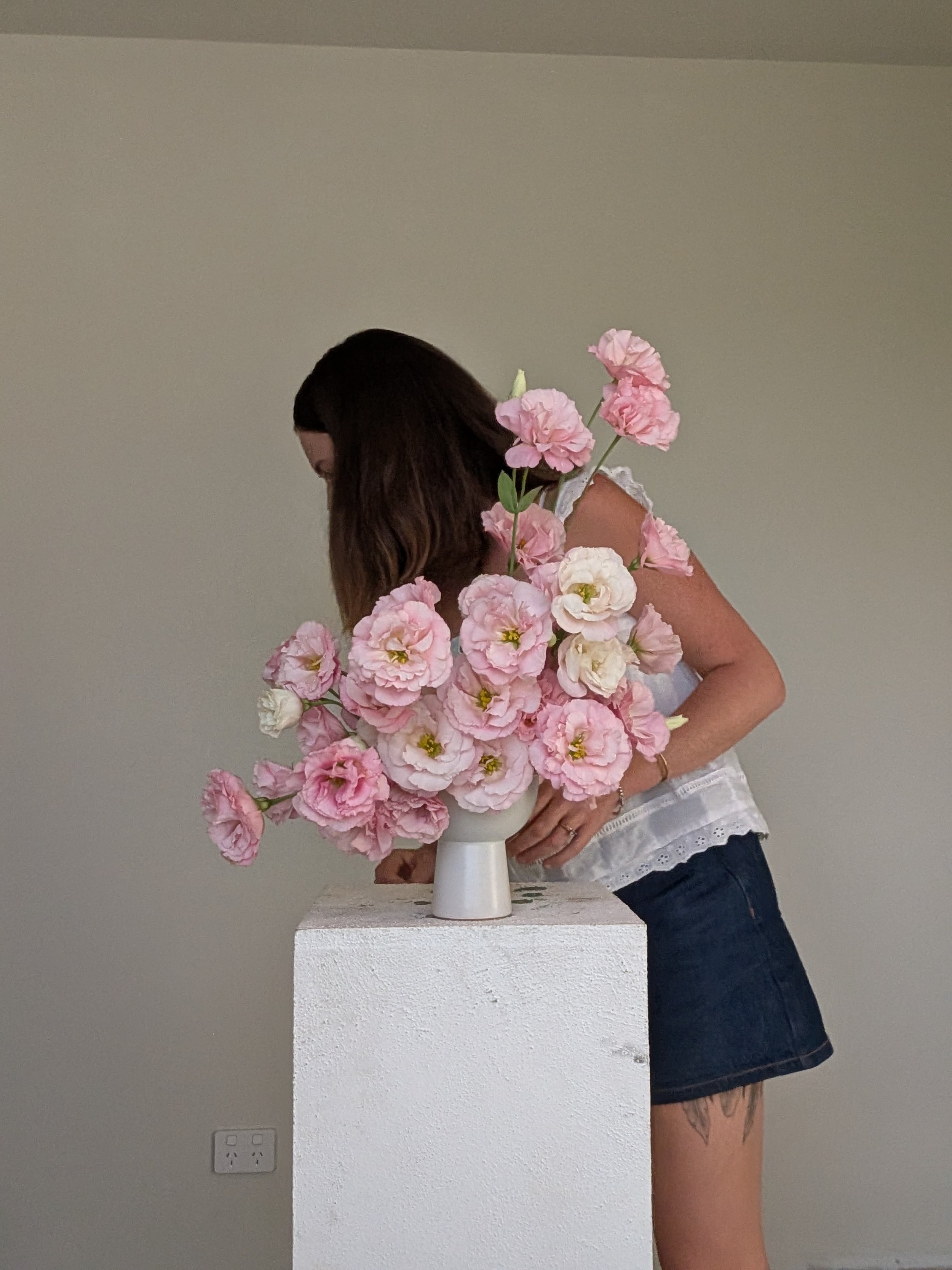 A woman with long brown hair in a white sleeveless top and denim shorts is standing next to a white table with a large bouquet of pink and white flowers in a white vase.