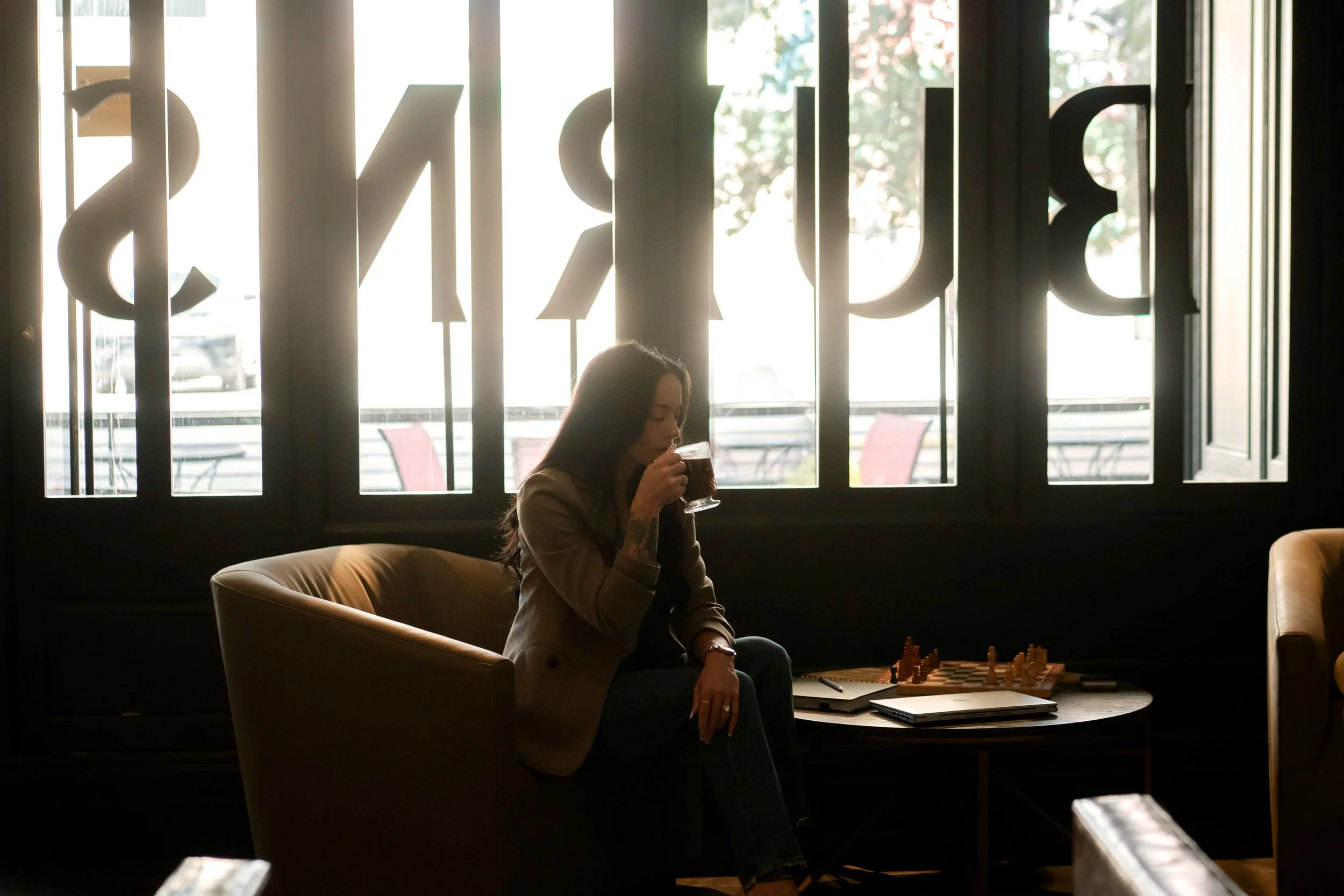 Gwen Taniguchi seated by a sunlit window, reflecting over a drink and a chessboard.