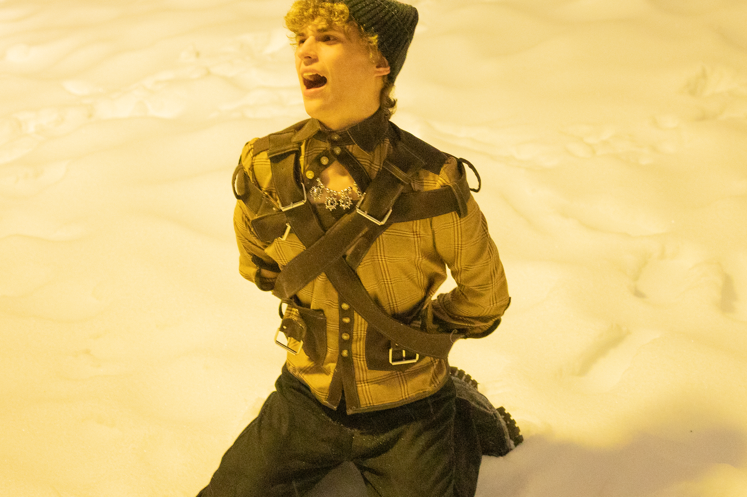 A person with curly hair wearing a black beanie, a plaid shirt, and black pants, kneeling on snow with a yellowish background, appearing to shout or scream.