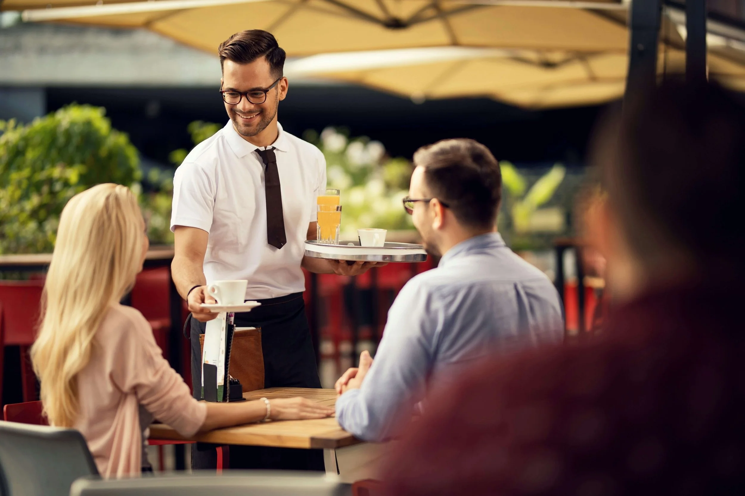 A male waiter in glasses and a white shirt with a black tie serves drinks to a group of customers seated at an outdoor restaurant table under large yellow umbrellas.
