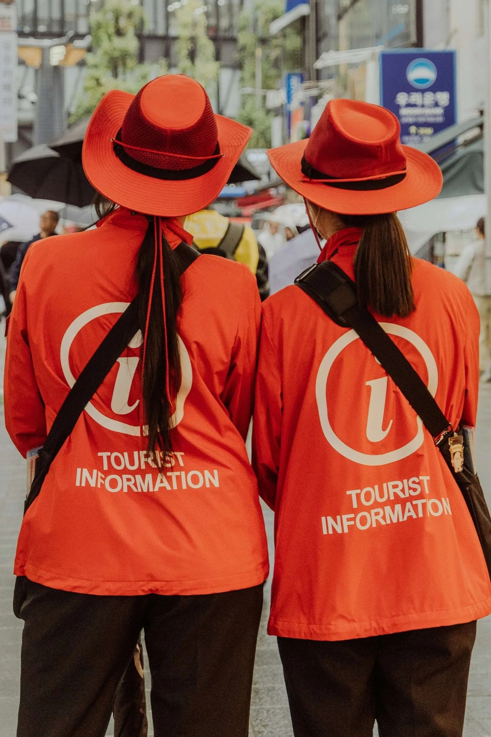 Two women wearing red tour guide uniforms and wide-brimmed hats, with the words 'Tourist Information' on their jackets, standing on a city street.