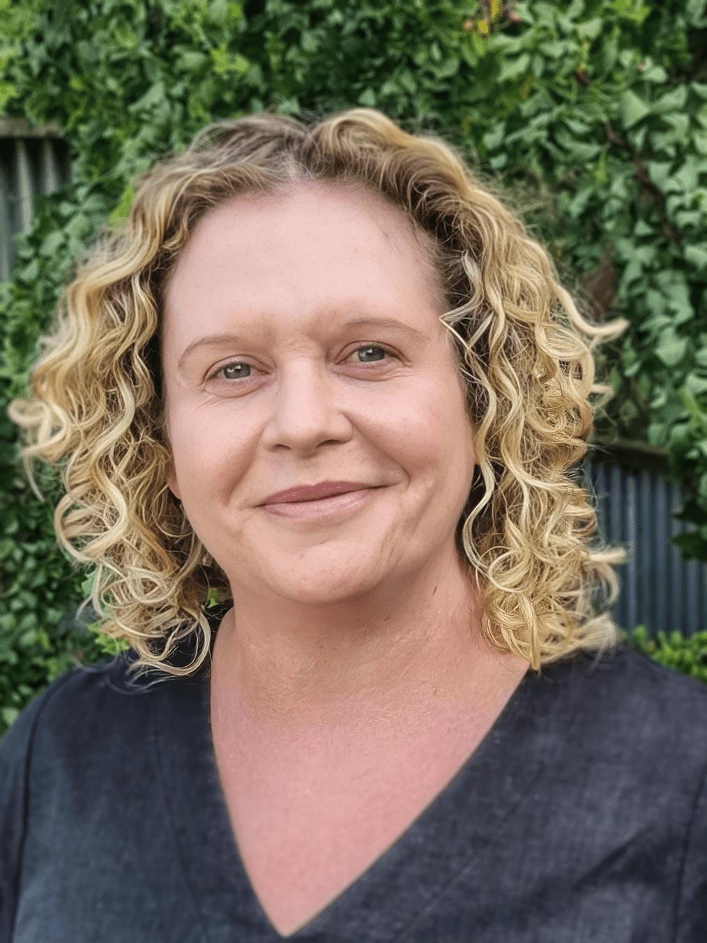 A woman with blonde, curly hair smiling outdoors in front of green foliage.