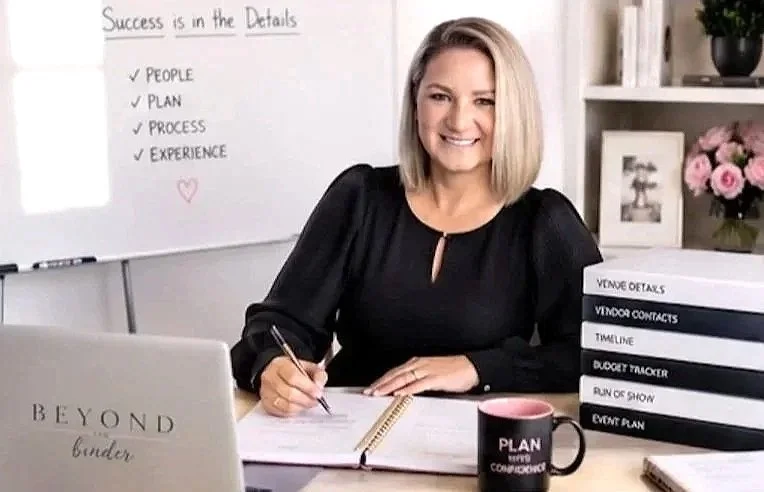A smiling woman sitting at a desk with a laptop, notebooks, and a pink coffee mug, in a bright office space with flowers and books.