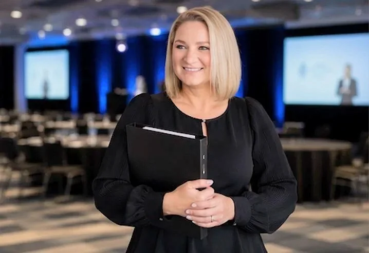 A woman in black holding a notebook at an indoor conference or presentation event with screens and empty chairs in the background.