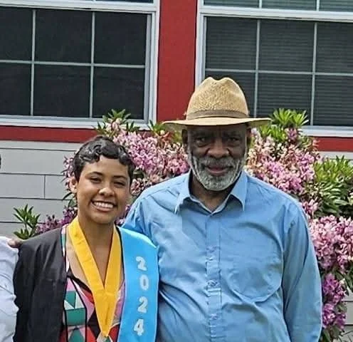 A young woman with short curly hair smiling, wearing a graduation gown and a blue sash with '2024' on it, standing next to an older man with a beard, gray hair, blue shirt, and straw hat, in front of pink flowering bushes and a building with large windows.