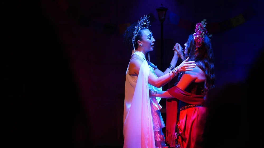 Two women dressed in elaborate costumes with headdresses are dancing together on a dark stage with dramatic lighting.