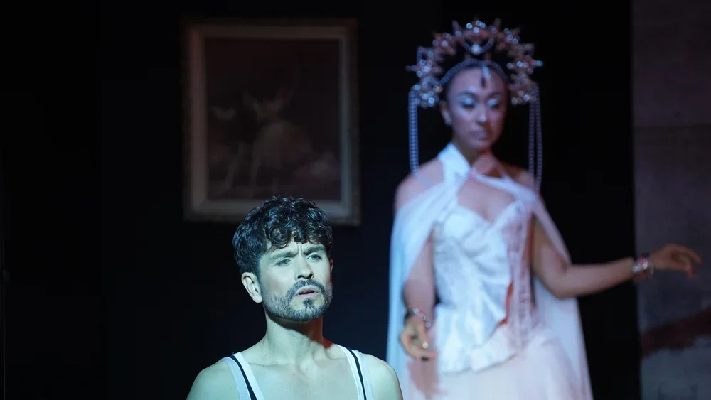 Two performers on stage, one in the foreground with a serious expression, the other in the background wearing an elaborate headdress and a white outfit, holding a pose.