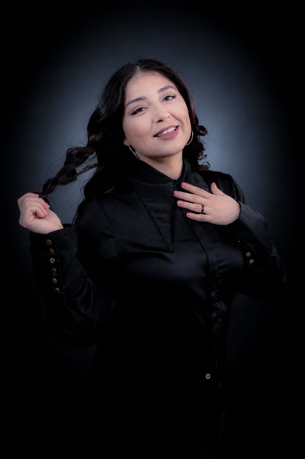 A young woman with dark, wavy hair, smiling, with one hand touching her chest and the other holding a braid, wearing a black jacket against a dark background.
