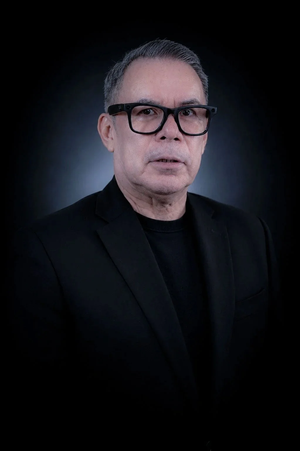 A portrait of an older man with gray hair, wearing black glasses and a black blazer, against a dark background.