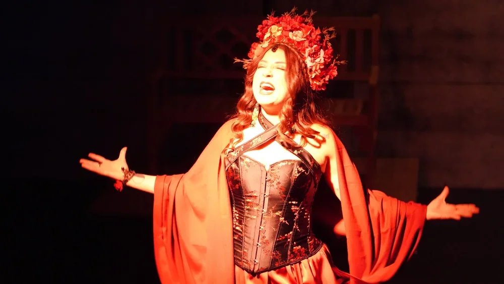 A woman performing on stage wearing a black corset, red cape, and a large floral headdress, with her arms outstretched and eyes closed.