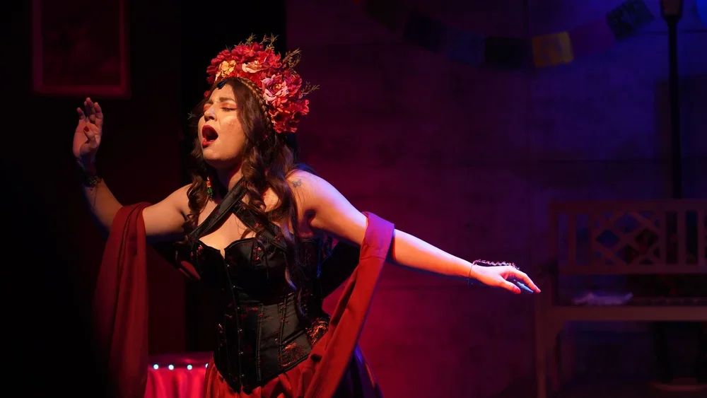 A woman wearing a floral headdress, black corset, and red shawl performing passionately on stage.