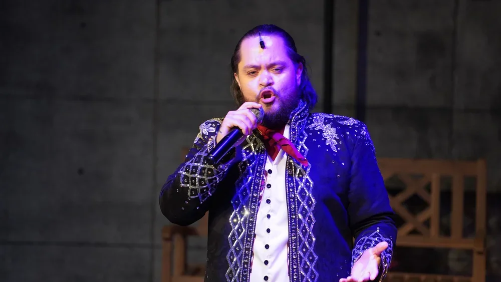 A man singing into a microphone, dressed in a decorated black jacket with silver embellishments, a white shirt, and a red scarf, against a gray background.