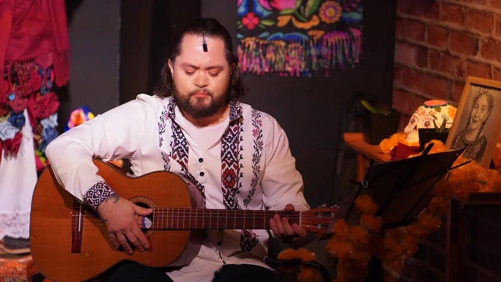 Man with long hair and beard playing an acoustic guitar, sitting in a decorated room with flowers and framed pictures.