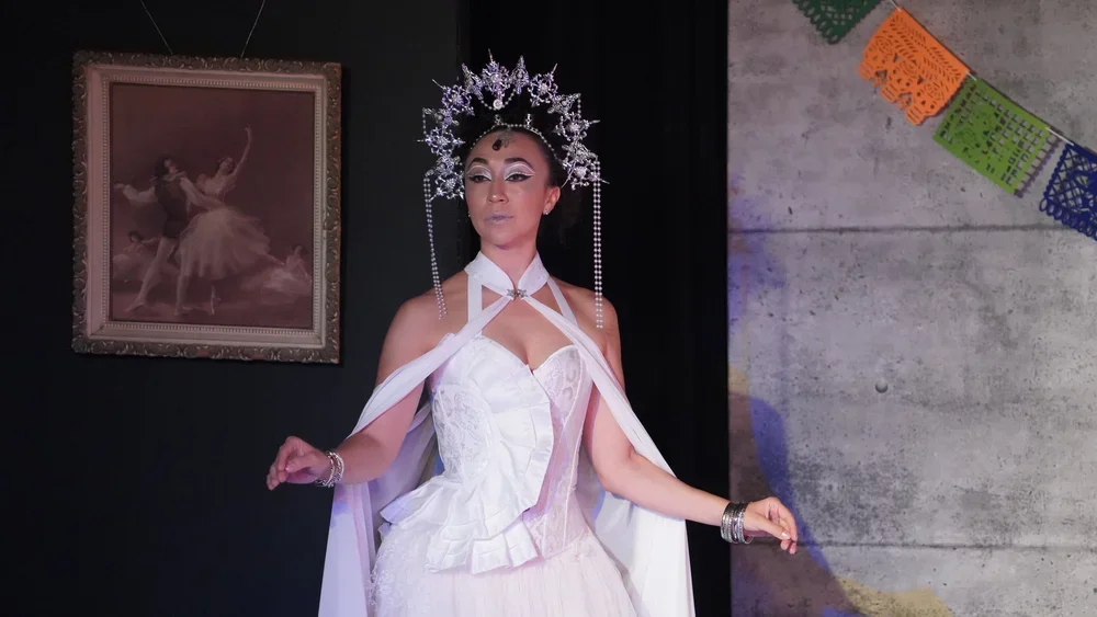 A woman dressed in a white gown with a cape, wearing a silver crown with pearl details, standing in front of a painting and colorful papel picado decorations.