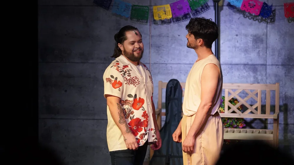 Two men standing face to face, smiling at each other, in front of a gray wall with colorful papel picado decoration above. One man has long hair and a beard, wearing a floral shirt, and the other has short hair and glasses, wearing a sleeveless beige
