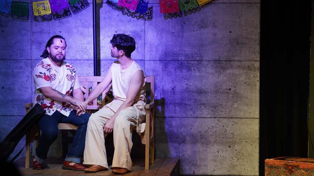Two men sitting on a wooden bench with dark concrete wall background, holding hands, in front of colorful papel picado banners.