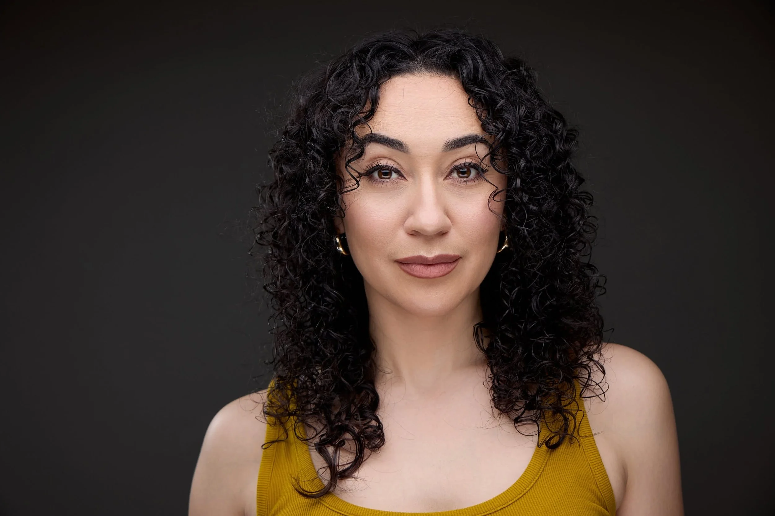 Portrait of a young woman with curly dark hair, wearing a yellow top, and smiling softly against a dark background.