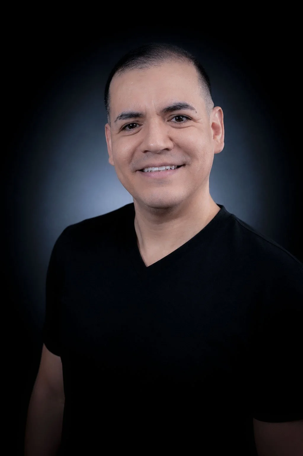 Portrait of a smiling man with short dark hair wearing a black V-neck shirt against a dark blurred background.