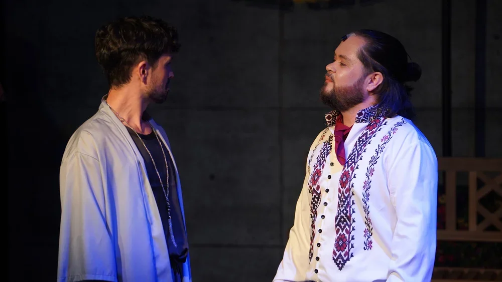 Two men face each other, one with dark hair and beard wearing a casual jacket, and the other with glasses, beard, long dark hair tied back, wearing a white embroidered shirt.