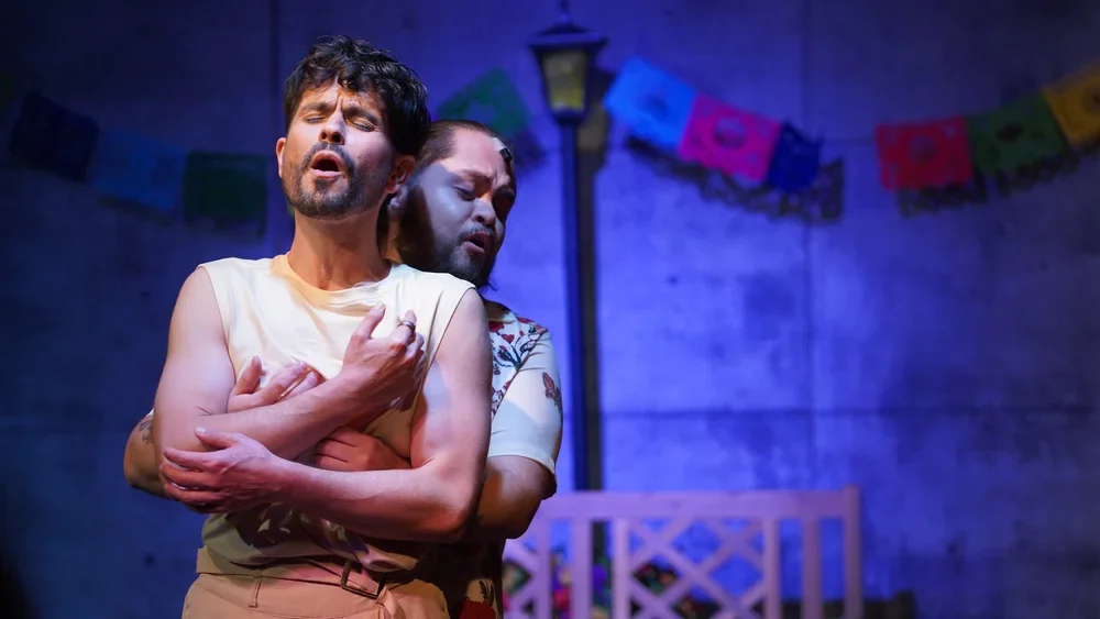 Two men are on stage, embracing each other with emotional expressions. The scene appears to be a theatrical performance set against a backdrop with colorful decorations and a streetlamp.