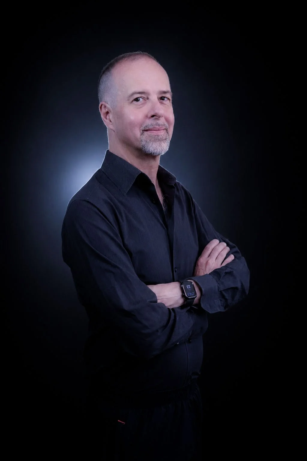 A middle-aged man with short hair, a goatee, and a confident expression, wearing a black shirt and smartwatch, standing with arms crossed against a dark background with a subtle light behind.