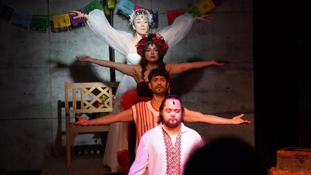 Four performers in traditional Mexican costumes pose with arms extended, creating a line against a plain wall decorated with colorful papel picado banners.
