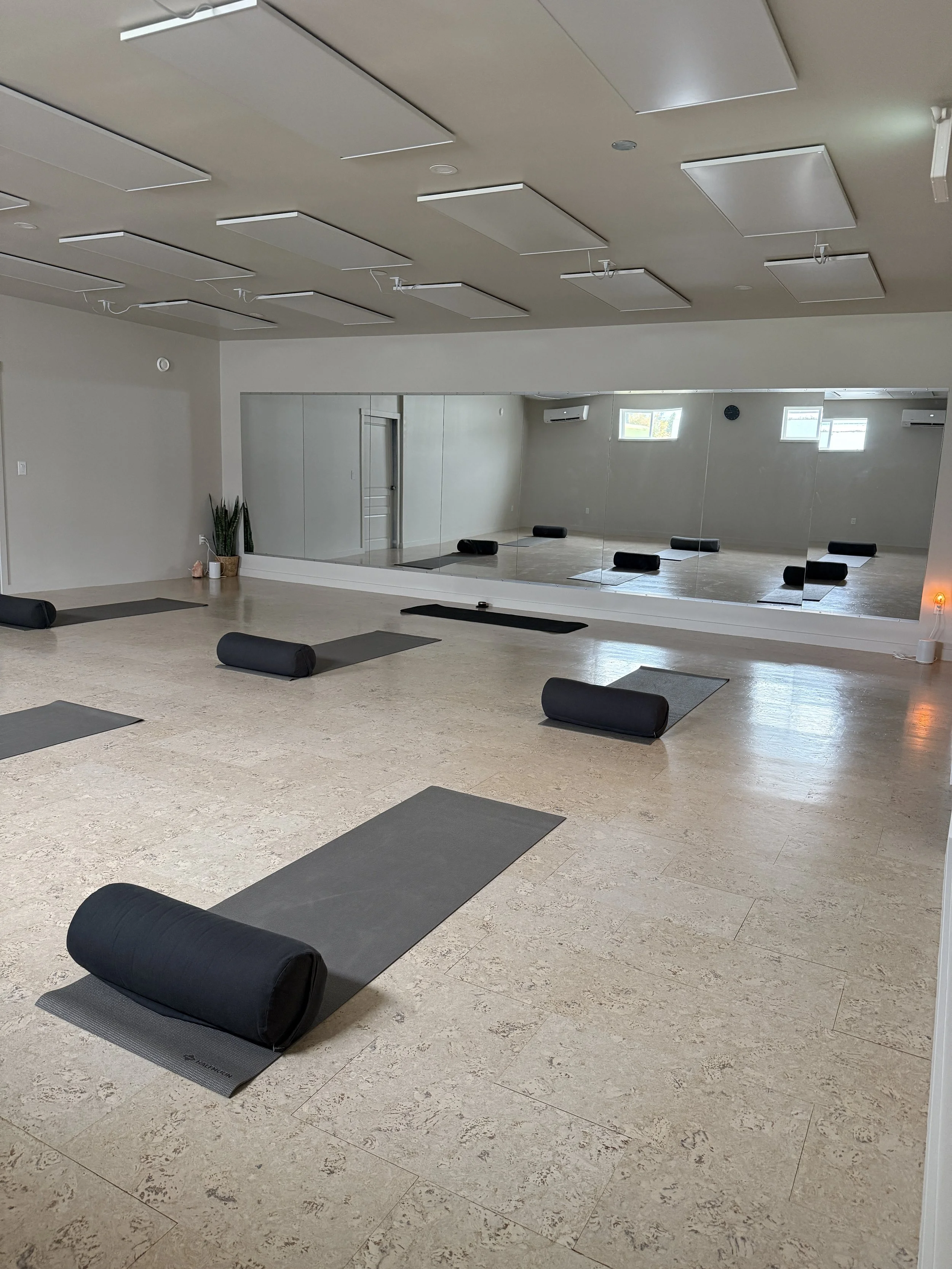 An empty yoga or meditation studio with mats and bolsters arranged on the floor, a mirror wall, and ceiling panels.