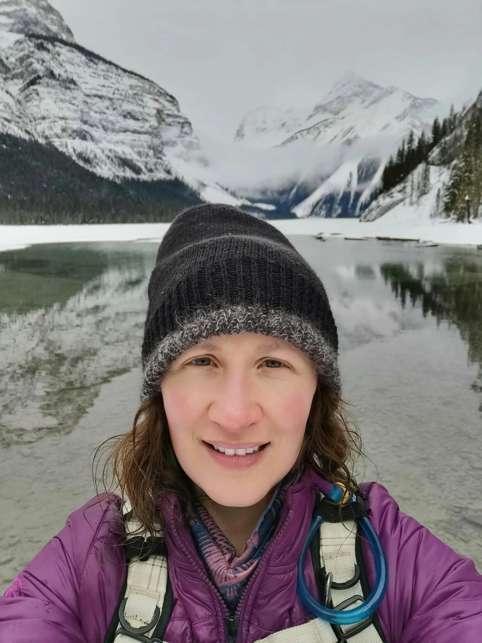A woman with brown hair, wearing a black knit hat, purple jacket, and a hiking backpack, smiling in front of a snow-covered lake with mountains in the background.