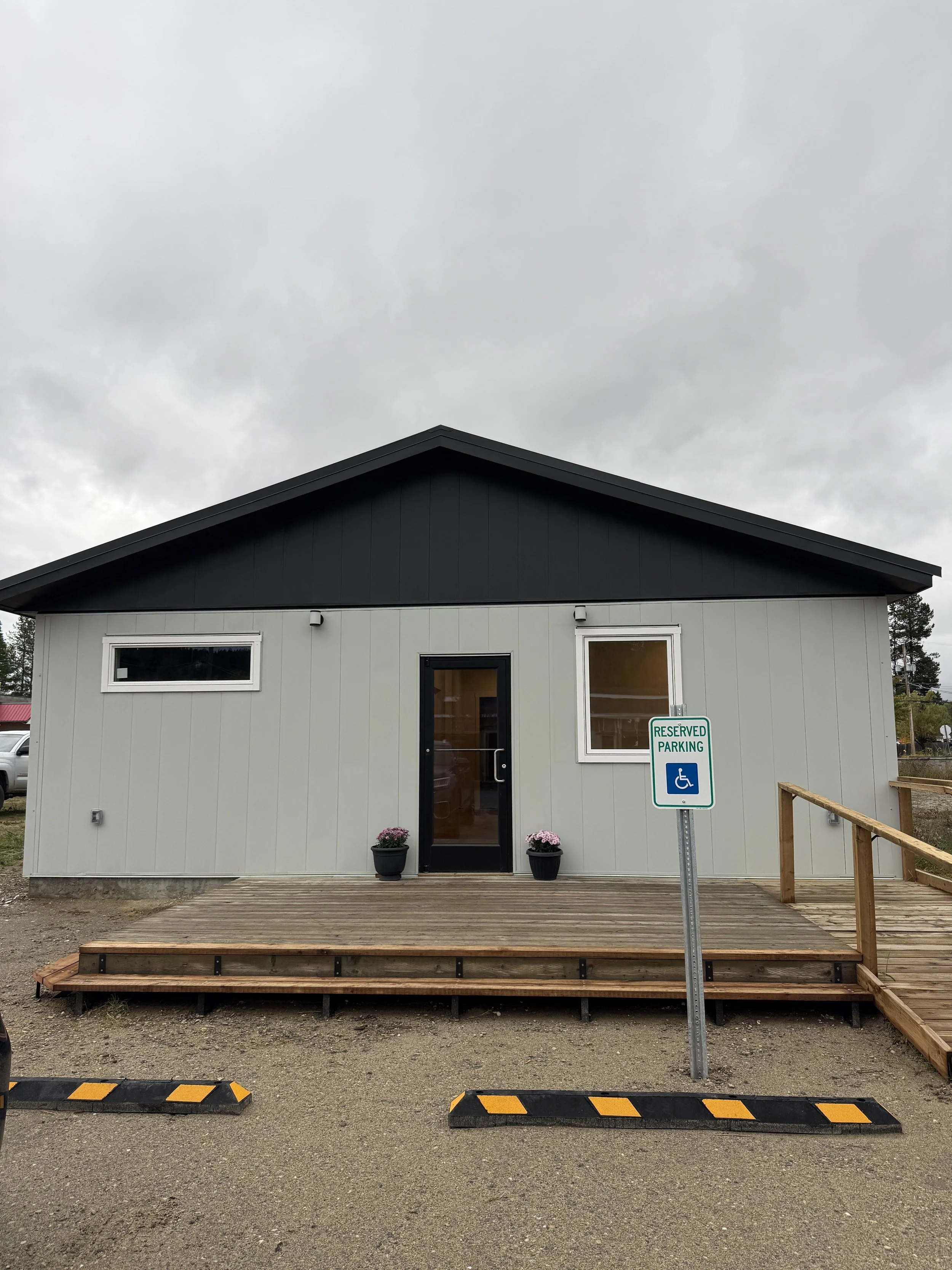 New small building with a black roof, gray exterior, large windows, and a locked door, with two flower pots with pink flowers at the entrance. A reserved parking sign with wheelchair accessibility is in front, and the sky is overcast.